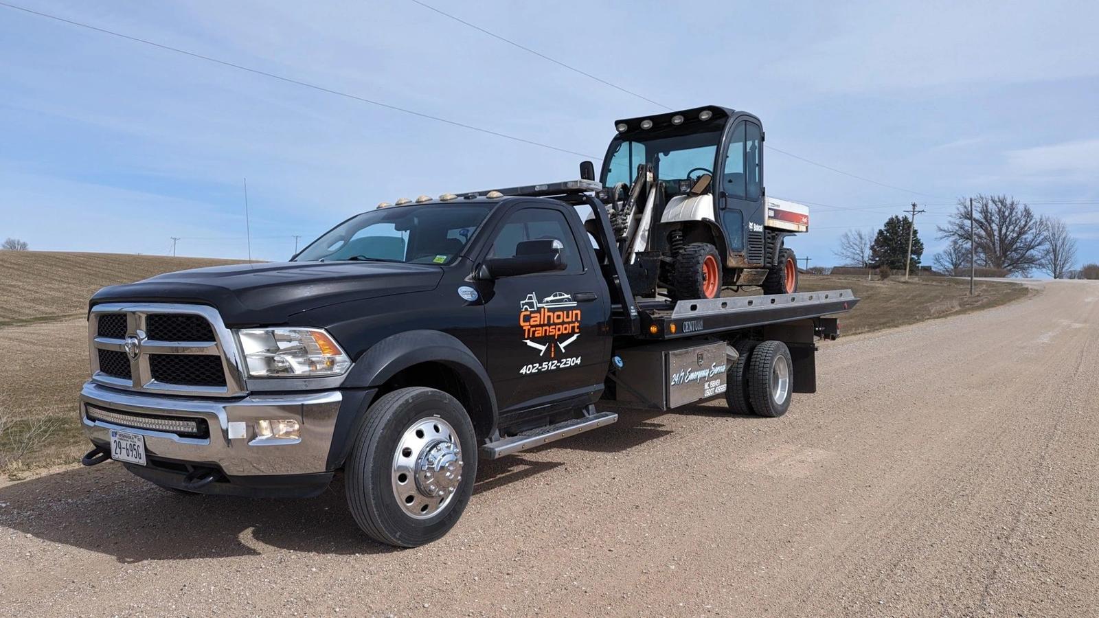 Calhoun Transport flatbed hauling a Bobcat skid steer on a gravel road in rural Nebraska