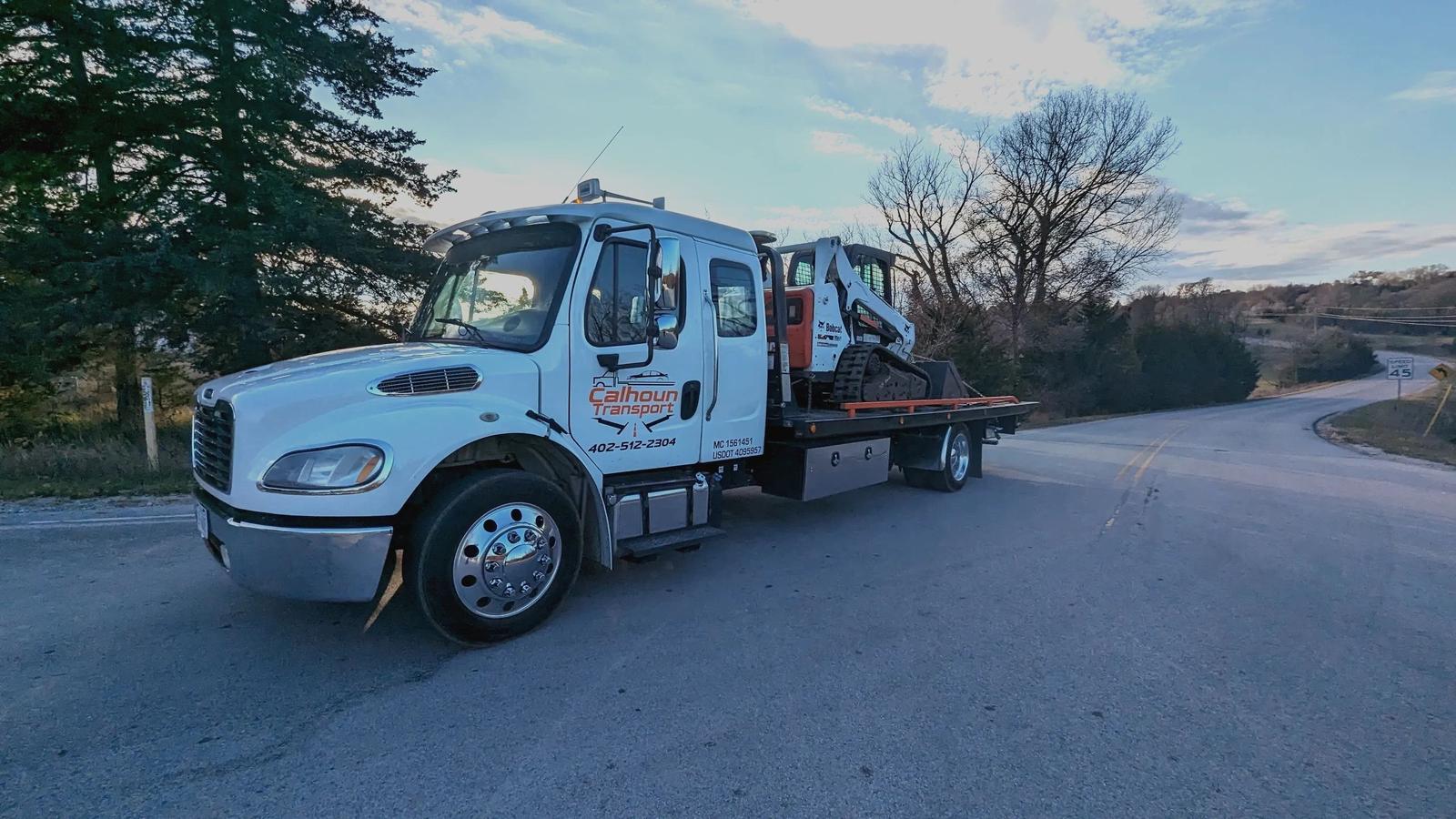 Calhoun Transport flatbed truck carrying a Bobcat skid steer on a Nebraska road at golden hour