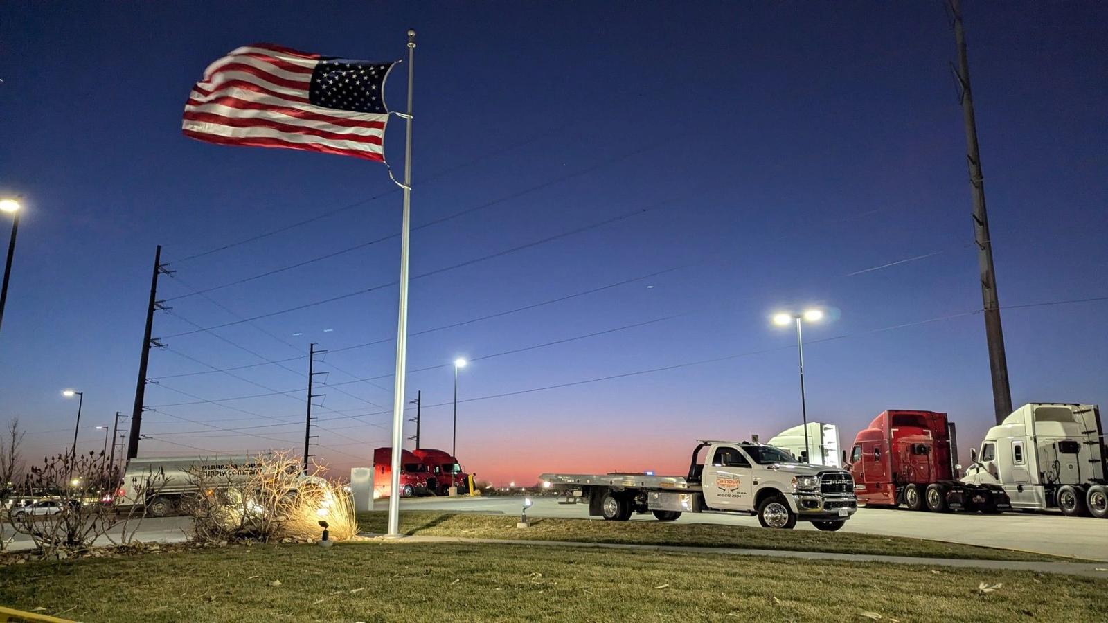 Calhoun Transport flatbed tow truck at a Nebraska truck stop beneath an American flag at dusk