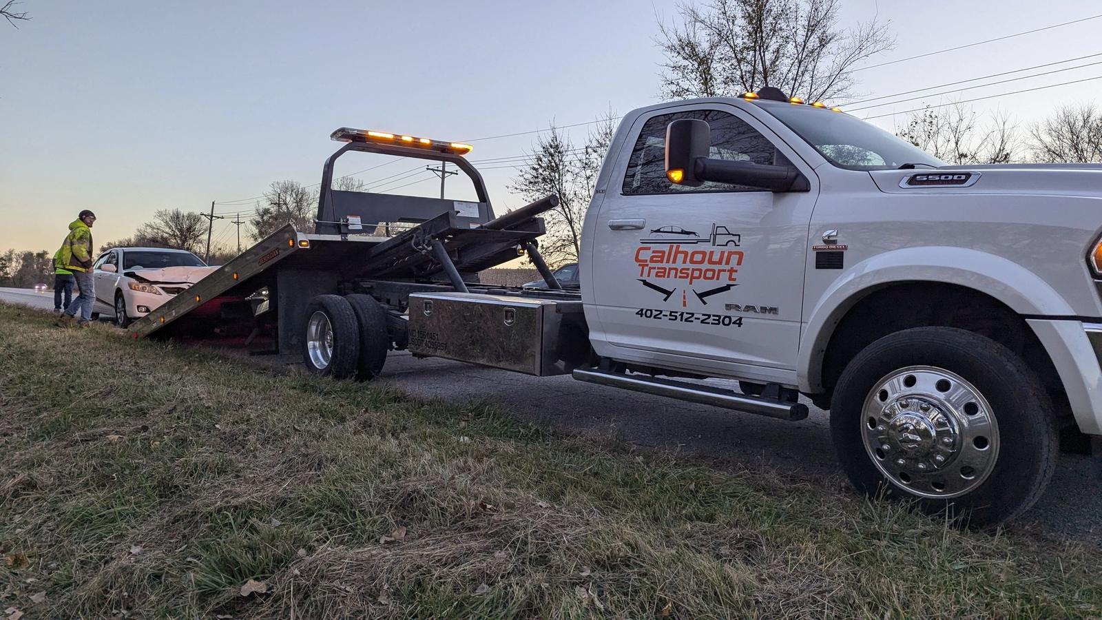 Calhoun Transport operator loading a disabled sedan onto a flatbed tow truck on a Nebraska roadside at dusk
